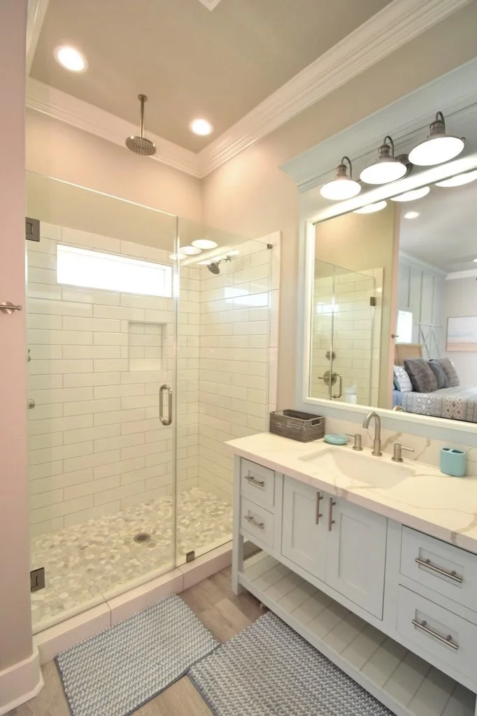 Bathroom with glass-enclosed shower, white subway tile, pebble floor, undermount sink, large mirror, vanity with drawers, and a bedroom visible through an open door.
