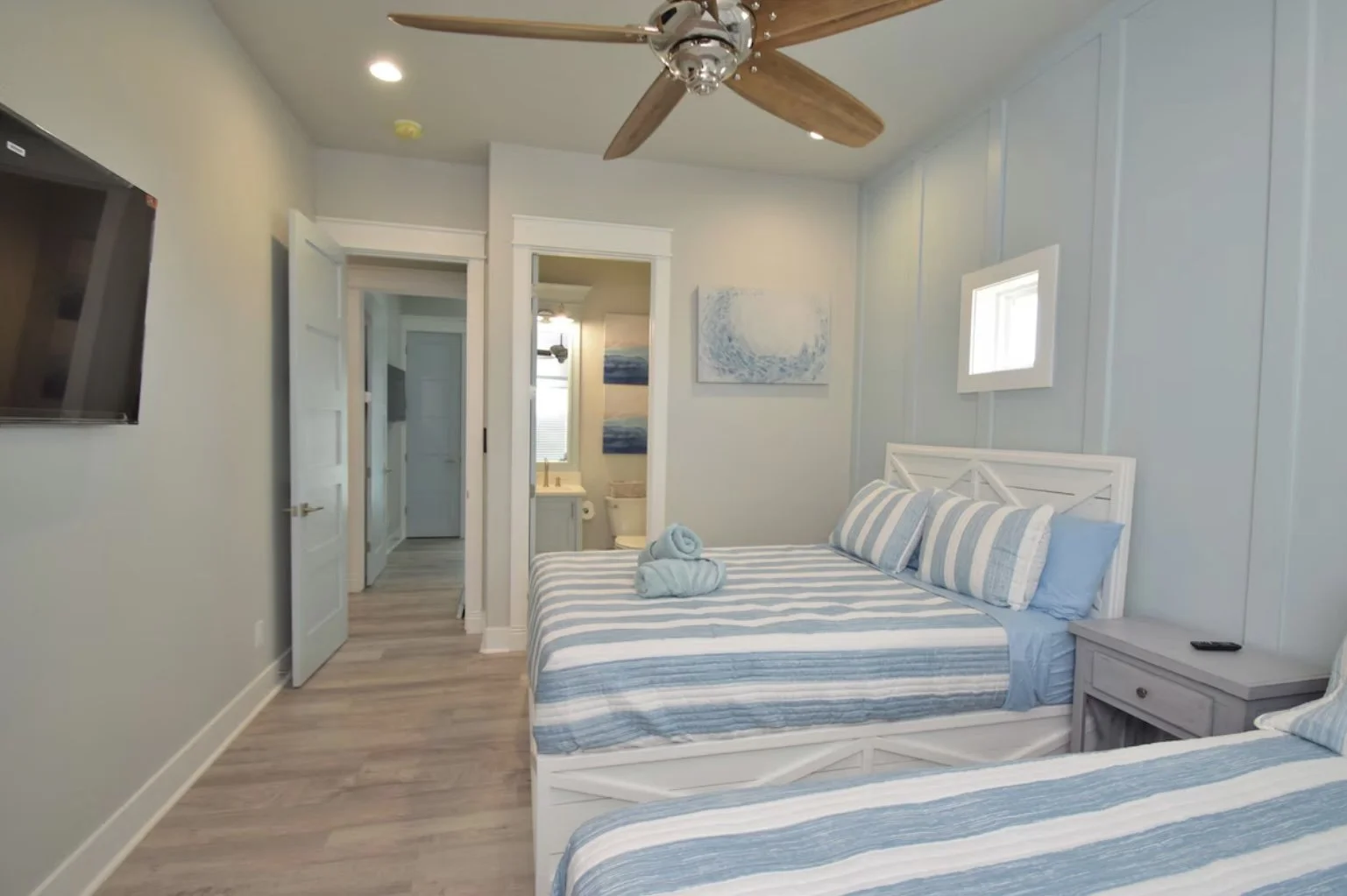 A bedroom with two beds featuring blue and white striped bedding, a small nightstand with a remote, wall art, and an entrance leading to a bathroom with a toilet and sink, a ceiling fan, and a mounted TV.