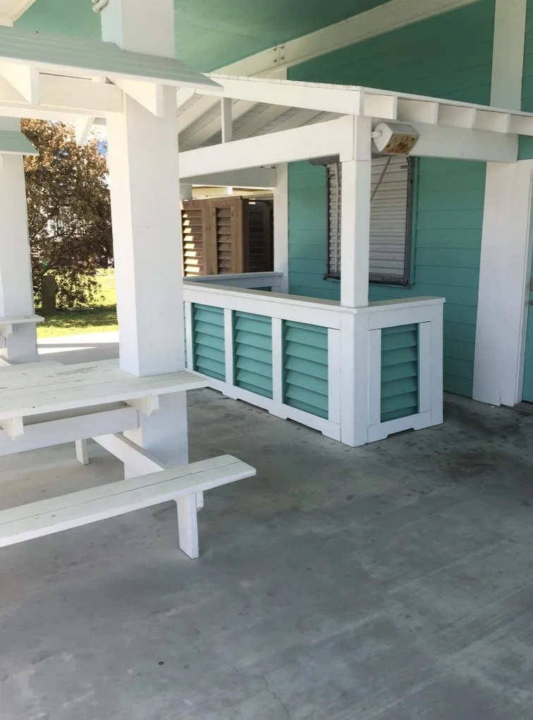 A white wooden picnic table on a concrete floor next to a teal-colored house with white trim and window shutters. A small porch with a railing is also visible.