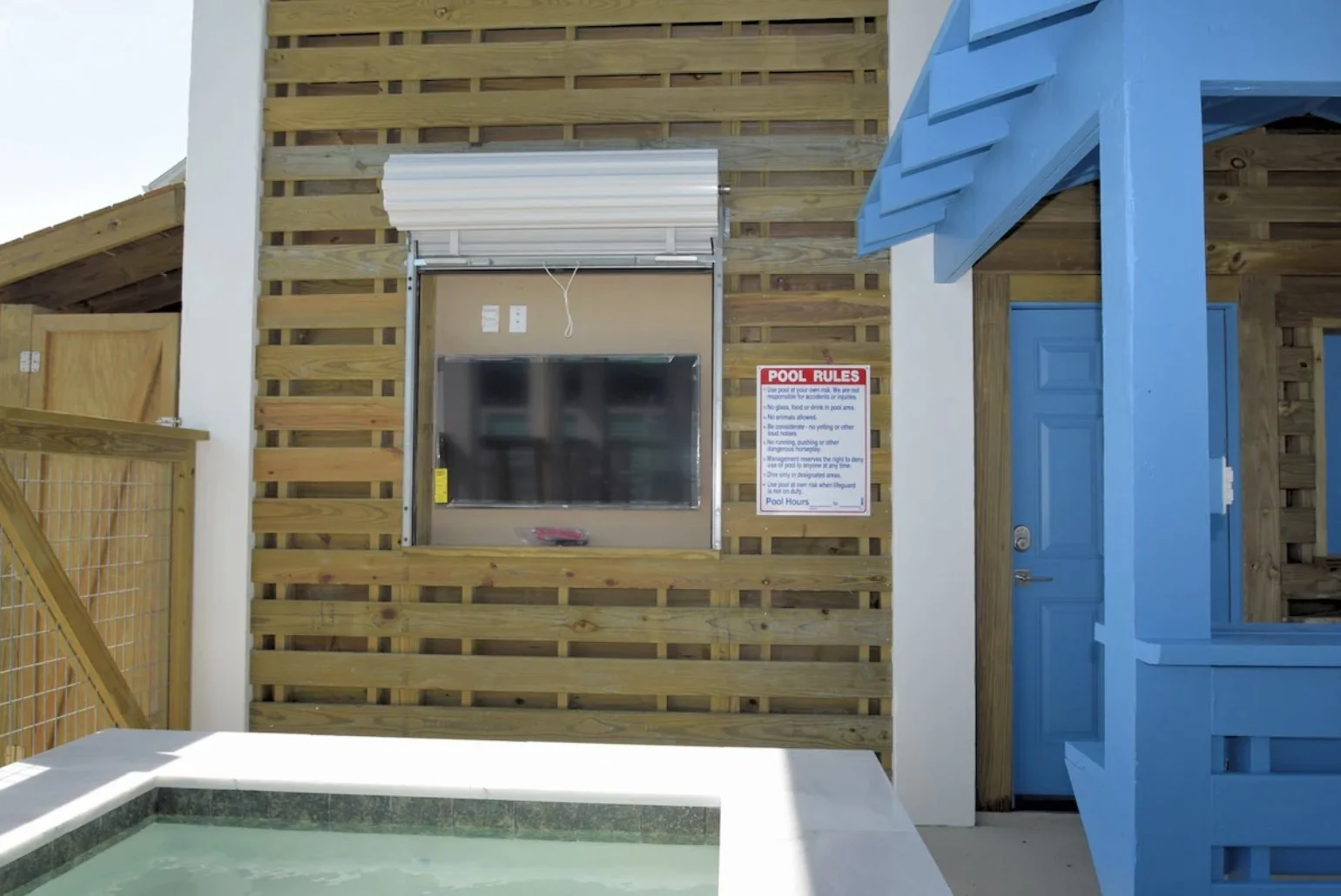 A poolside area featuring a wooden privacy wall, a mounted television under a metal roll-up cover, a sign listing pool rules, part of a hot tub in the foreground, a white wall, and a blue door with a small overhang.