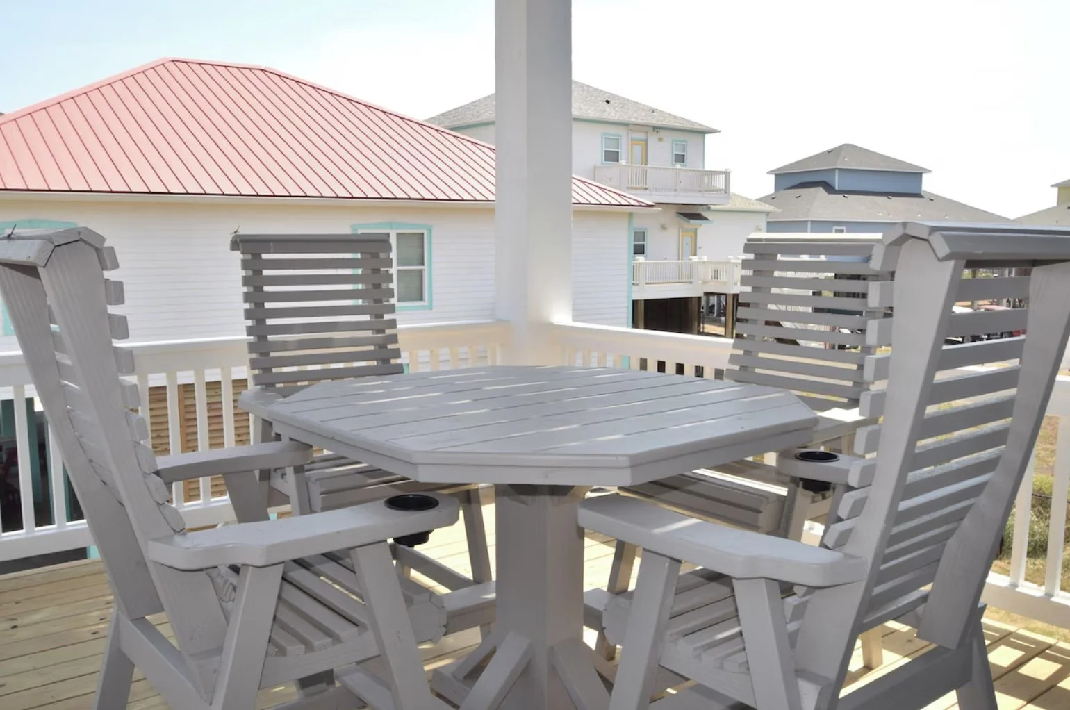 Outdoor balcony with a white round table and four white wooden chairs, overlooking neighboring houses with pastel-colored exteriors and balconies, under a clear sky