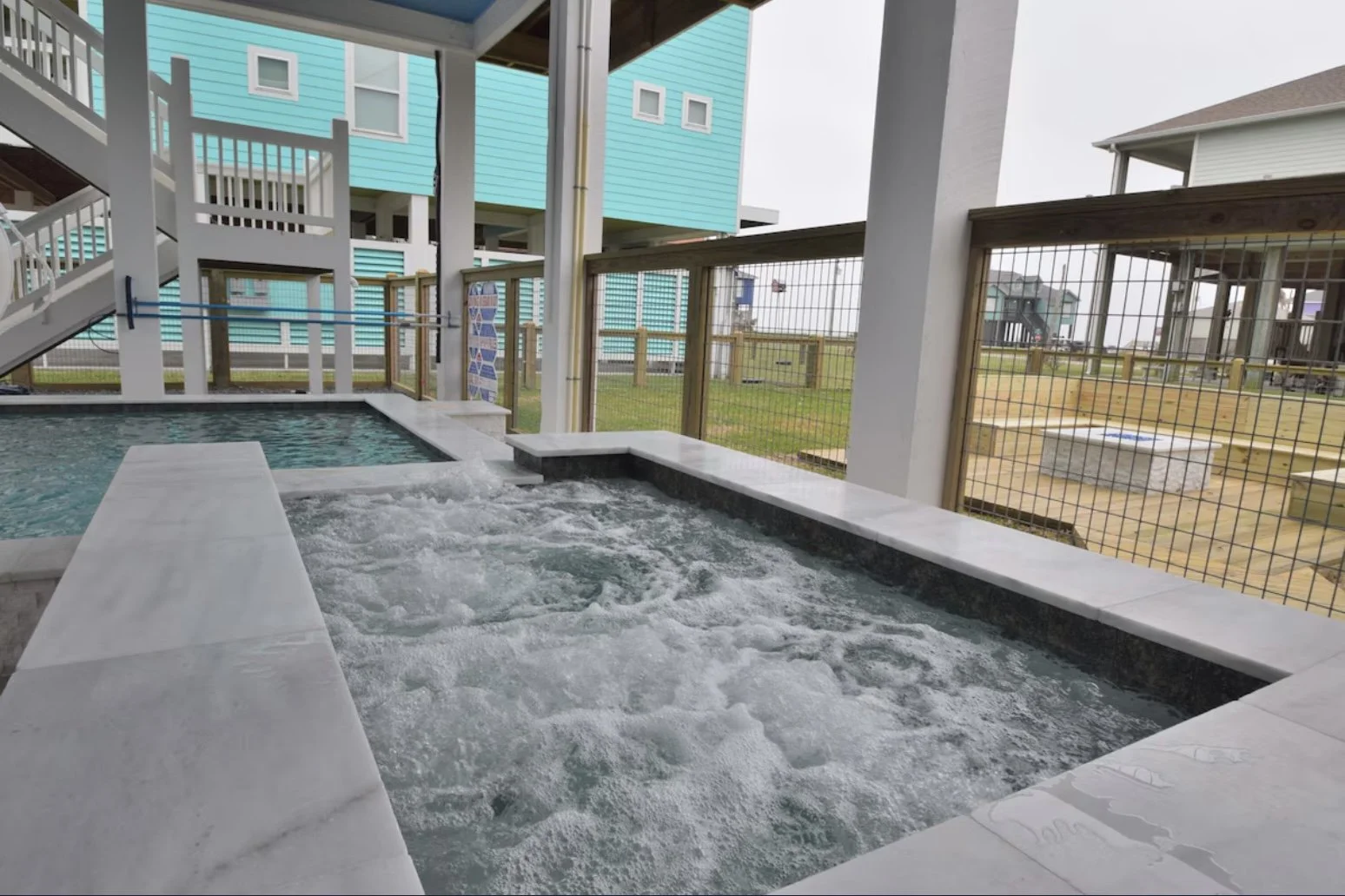 Outdoor hot tub with bubbling water, surrounded by a white marble edge, in a seaside resort area with beach houses in the background.