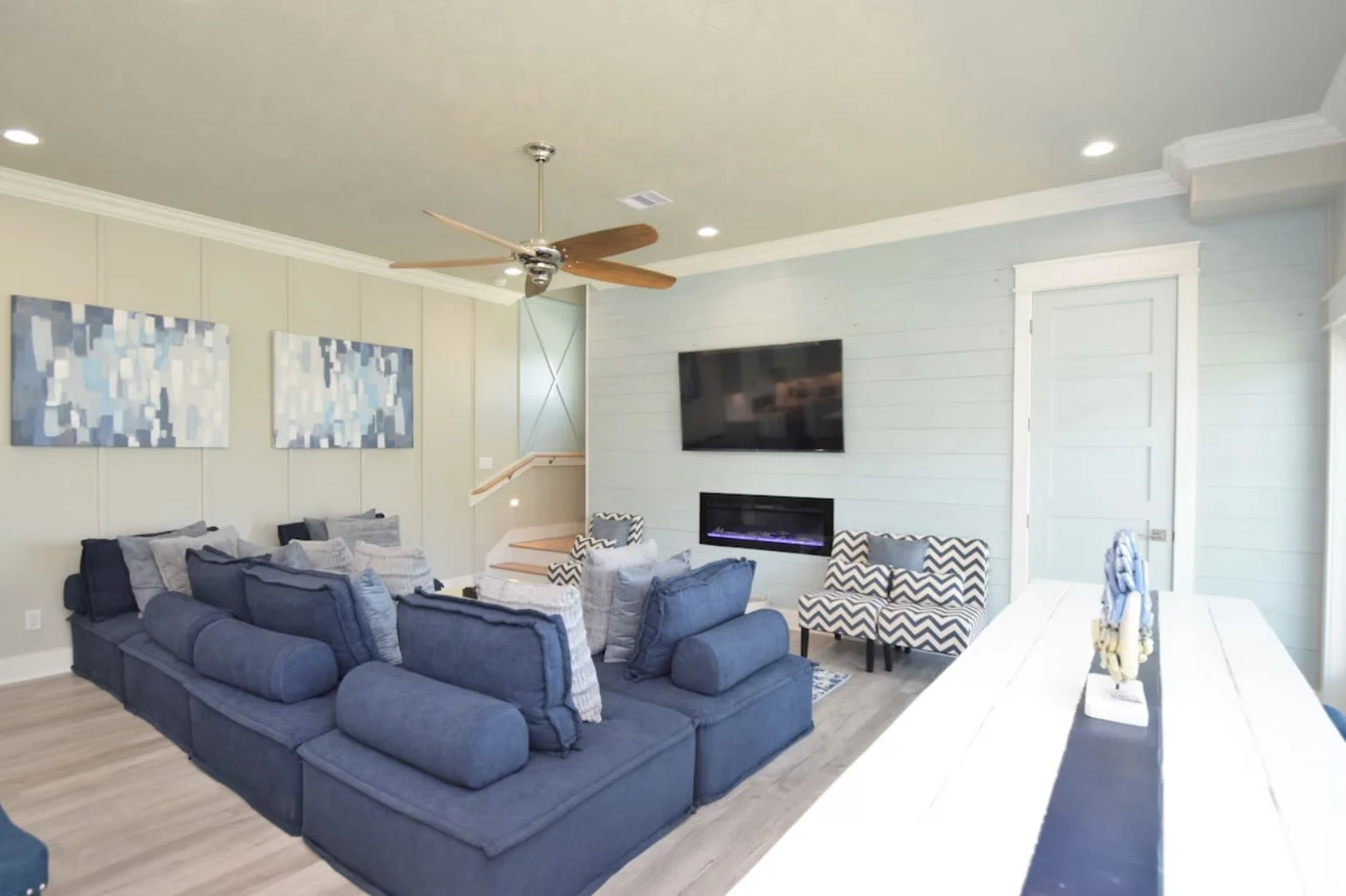Living room with a large blue sectional sofa, patterned armchairs, wall-mounted TV, modern electric fireplace, artwork, ceiling fan, and a dining table.
