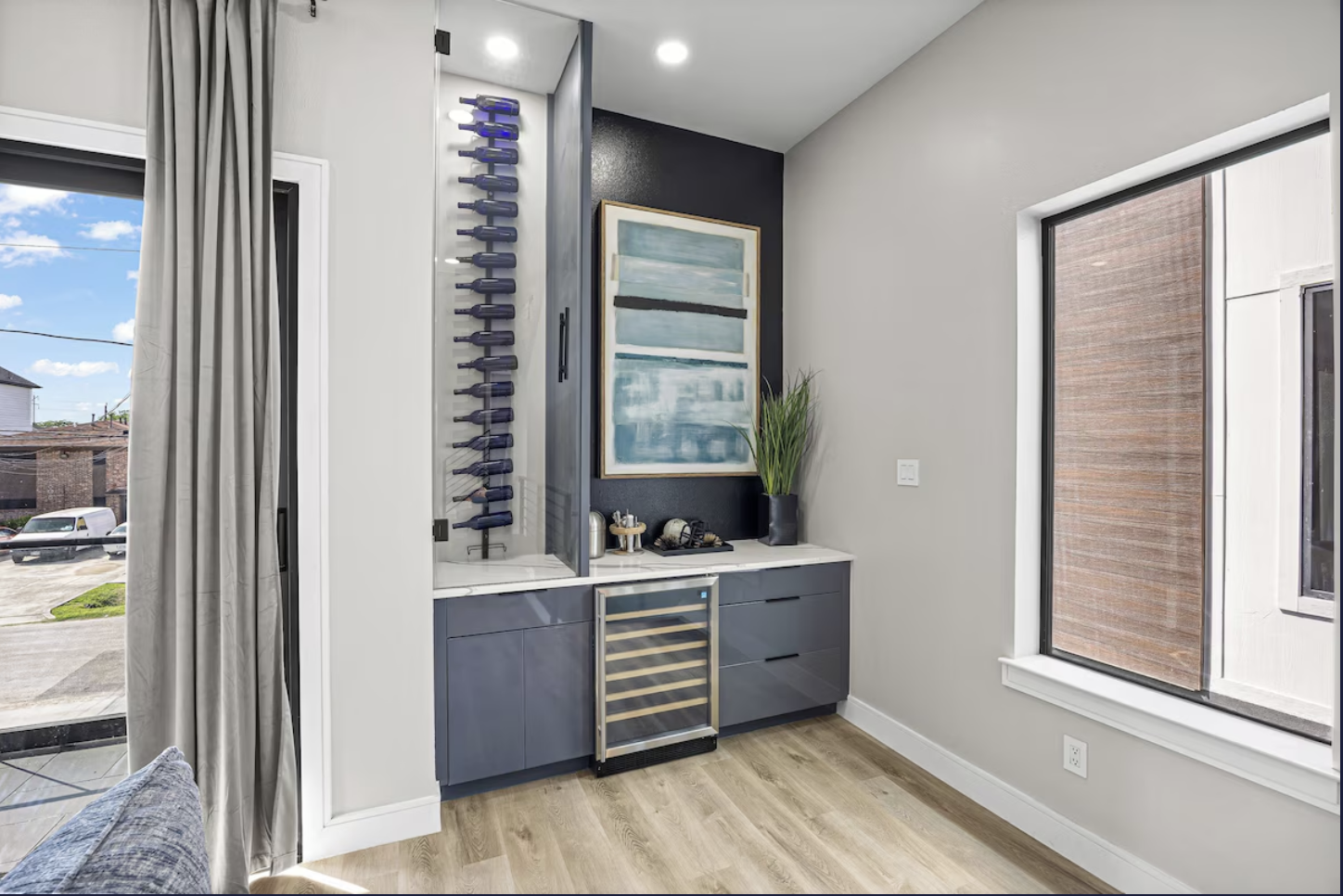 Interior of a modern room with a built-in mini bar, wine bottles, abstract artwork, a potted plant, window with fabric shades, and light wood flooring.
