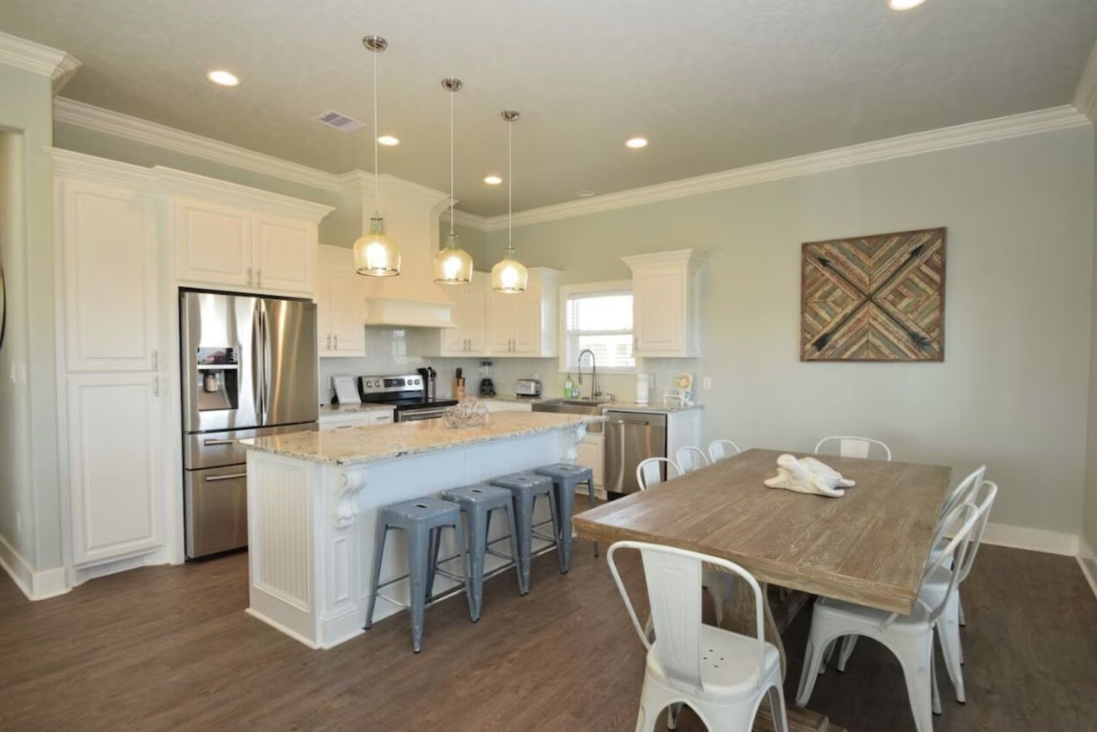 Kitchen with white cabinets, stainless steel refrigerator, granite countertop island, four gray bar stools, wooden dining table with white chairs, pendant lights, and wall art.