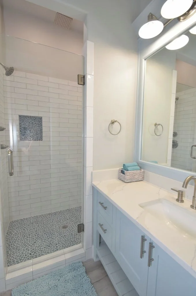 Bathroom featuring a glass shower with white subway tiles, a pebble stone shower floor, a built-in shower niche, a white vanity with a marble countertop, a large mirror, and white walls.
