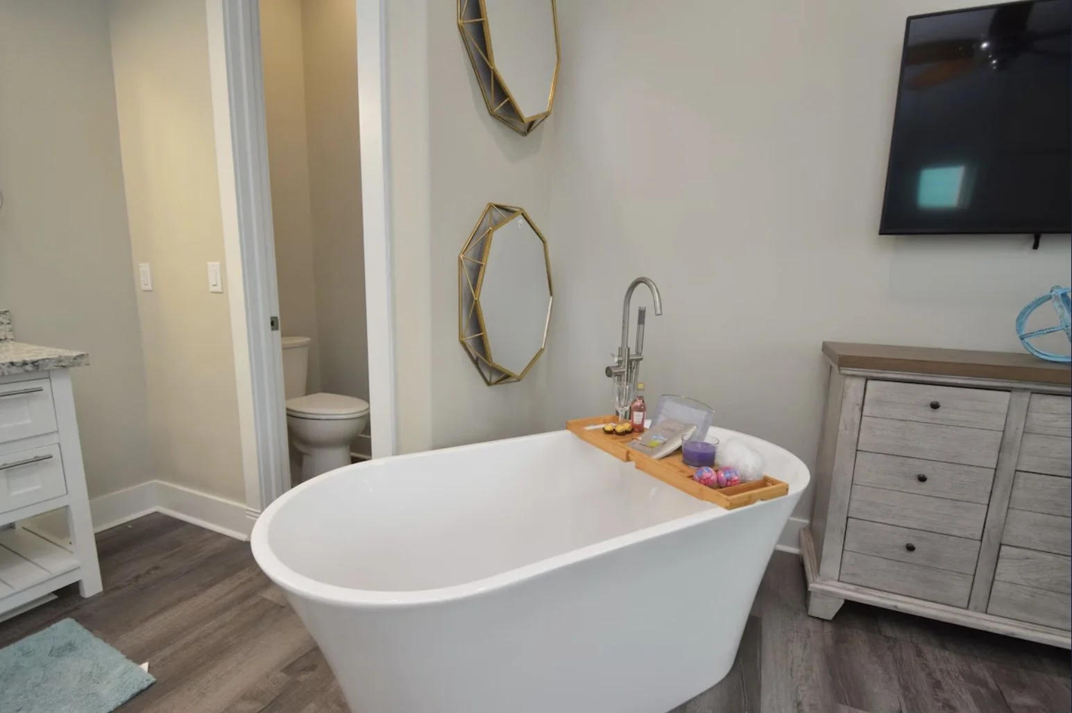 Bathroom with a freestanding bathtub, a wooden top tray with bath items, a wall mirror, and a closet. A separate toilet is visible through an open door.