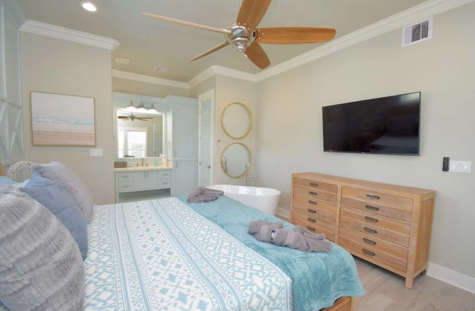 A bedroom with a bed, a TV mounted on the wall, a wooden dresser, a bathtub, and bathroom vanity in the background, with beige walls and ceiling fan.