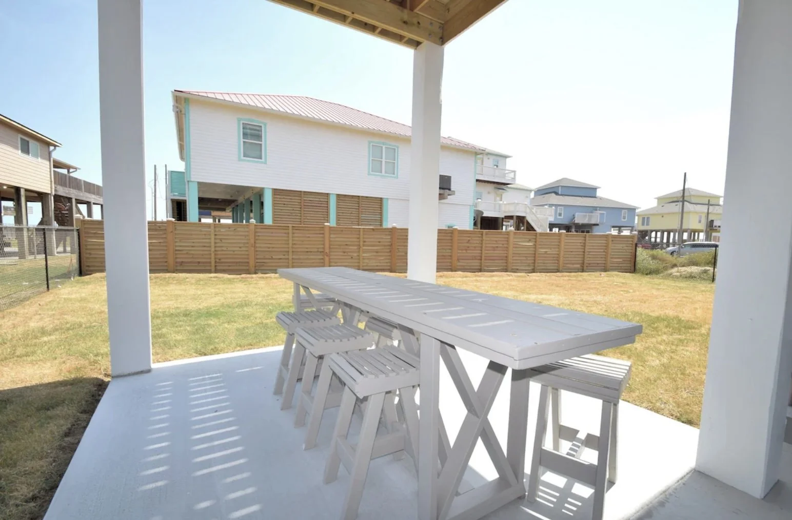View from a covered porch of a backyard with a white picnic table and stools, wooden fence, and neighboring colorful houses in a coastal area.