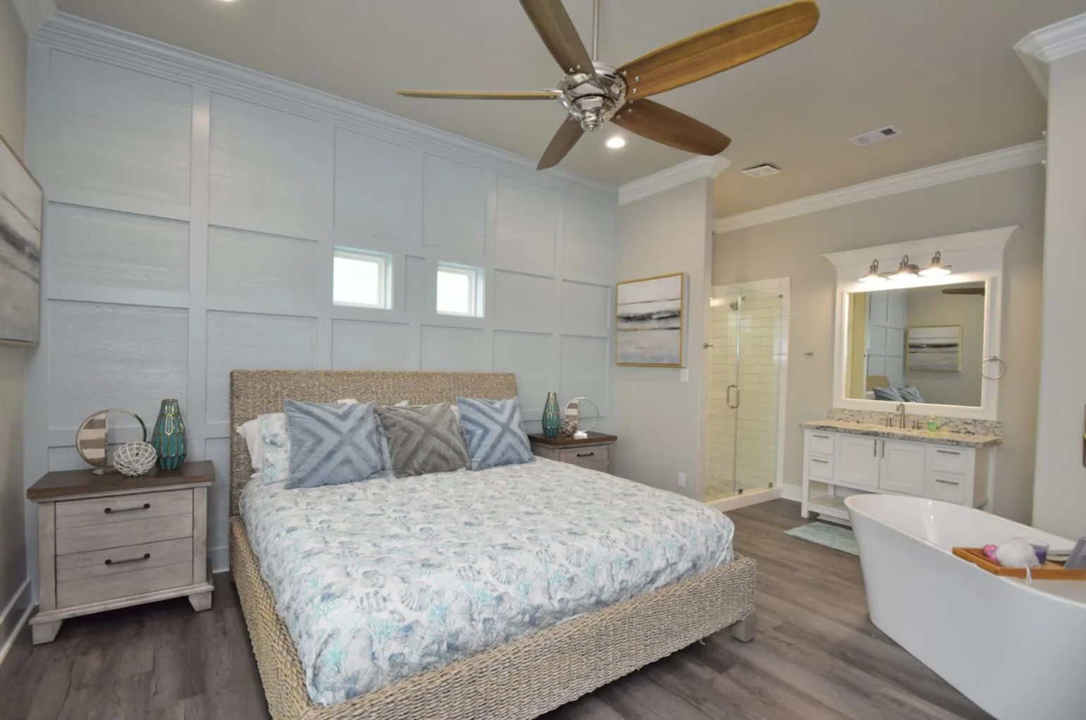 A modern bedroom with a large bed, nightstands, and decorative pillows. There's a walk-in shower and a vanity with a mirror, and a bathtub with bath toys, all in a neutral color scheme.