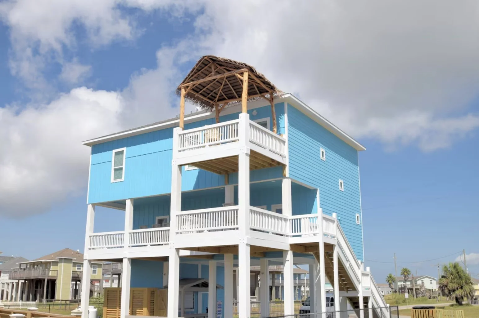A multi-story beach house painted in bright blue with white railings, elevated on stilts, with a thatched roof deck on the top level, against a partly cloudy sky.