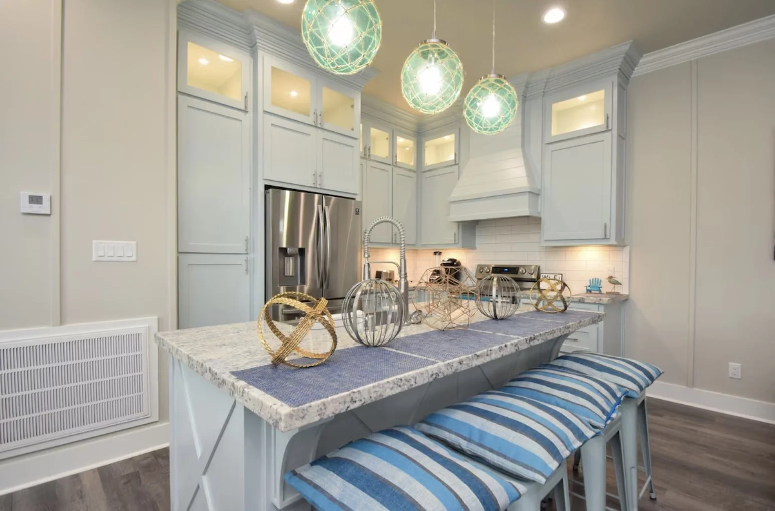 Modern kitchen with blue cabinetry, stainless steel refrigerator, granite island with blue runner, striped seating, and decorative lanterns hanging from ceiling.