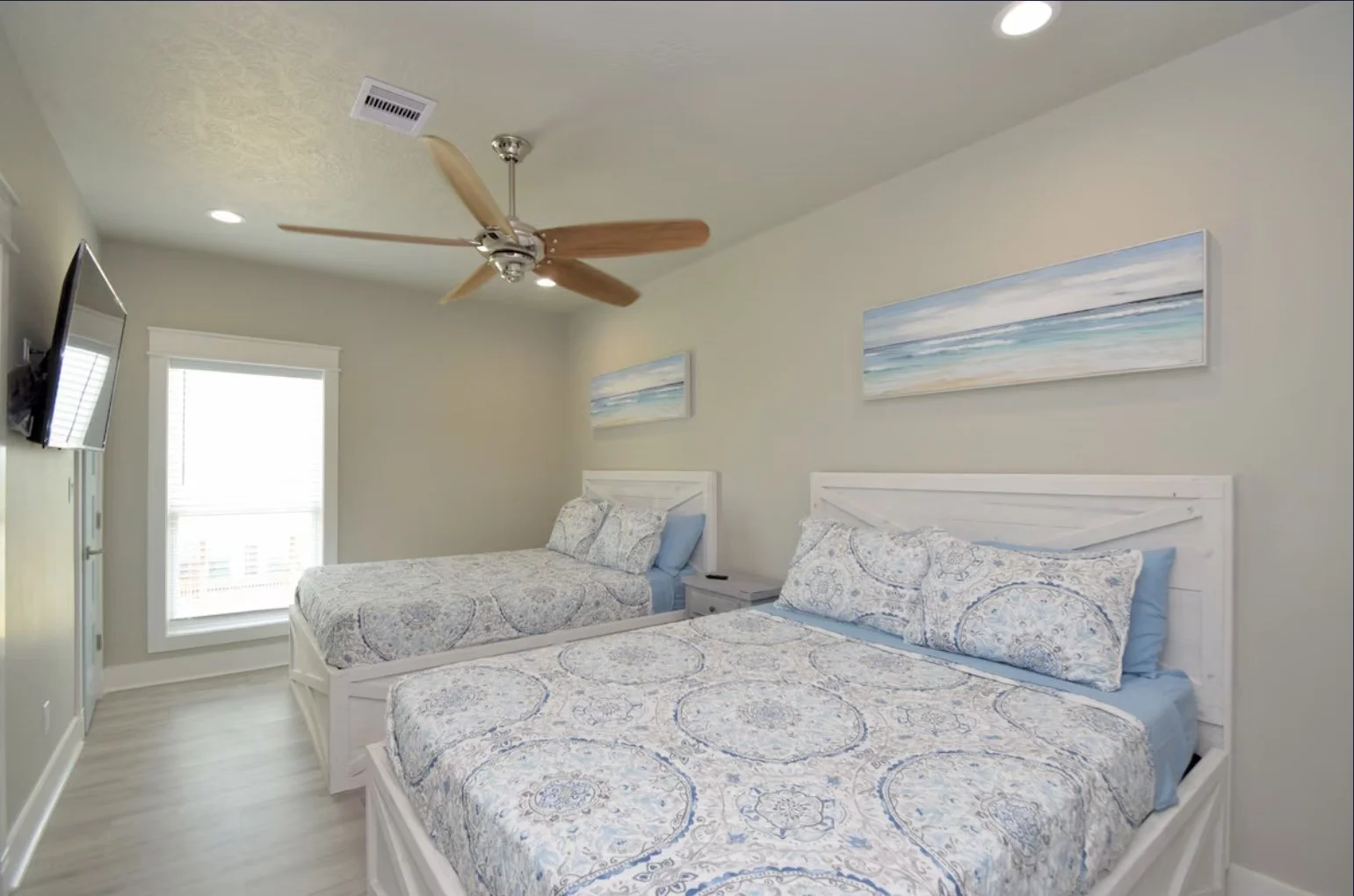 A bedroom with two white beds with blue and white patterned bedding, a nightstand, wall art of a seascape, a ceiling fan, and a window with blinds.