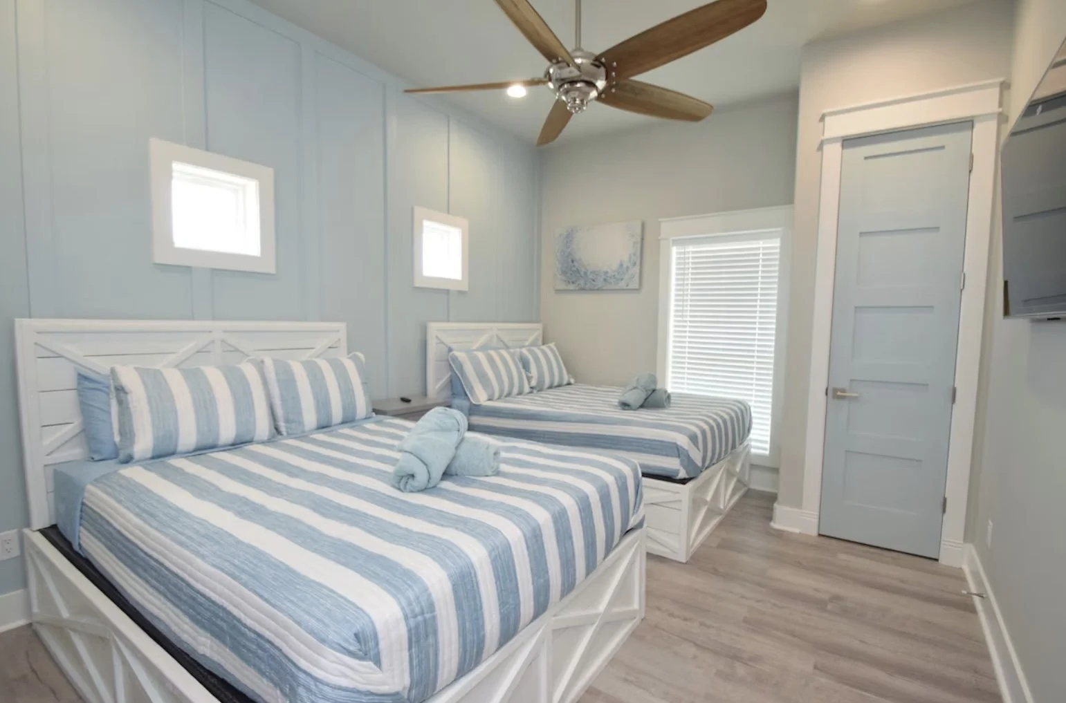 A bedroom with two twin beds featuring white wood frames and blue and white striped bedding, two rolled towels on each bed, two small windows above the beds, a ceiling fan, a door, a window with blinds, a wall-mounted TV, and a piece of wall art.