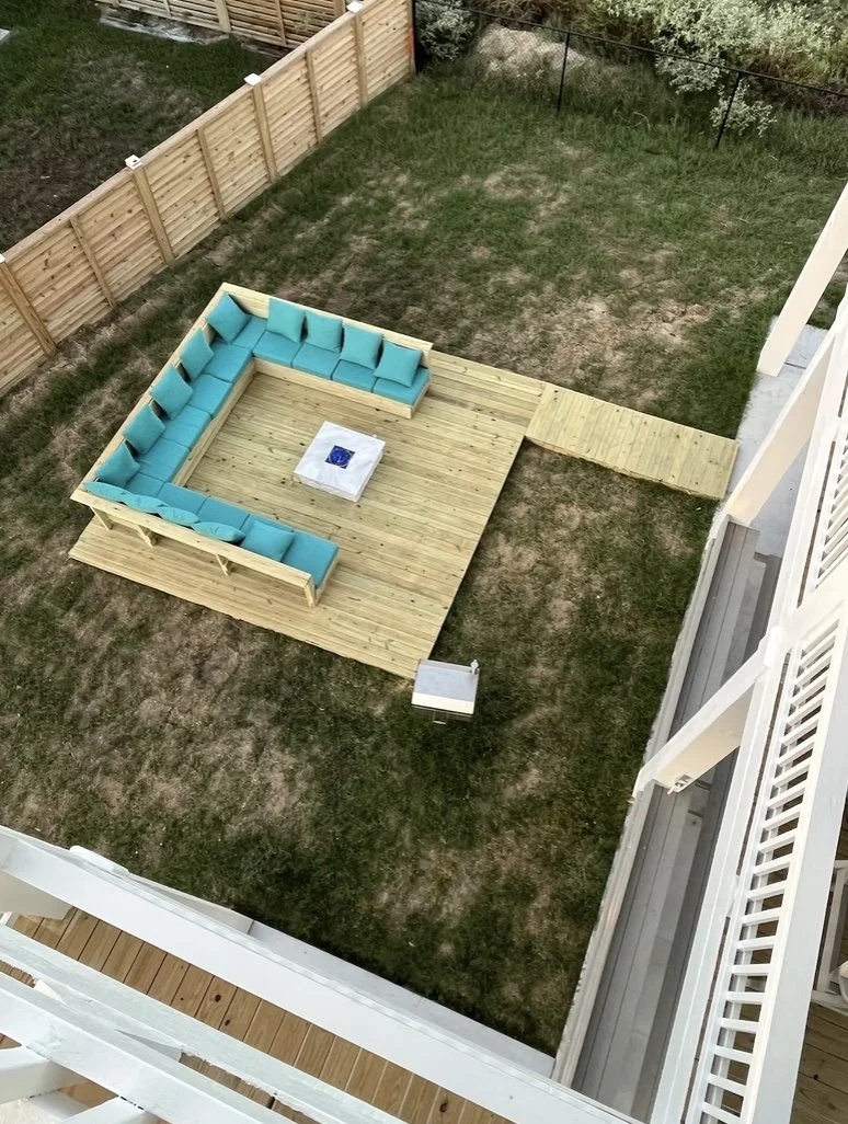 An aerial view of a backyard with a wooden deck and blue outdoor sectional sofa around a fire pit, surrounded by a grassy lawn and a wooden fence.