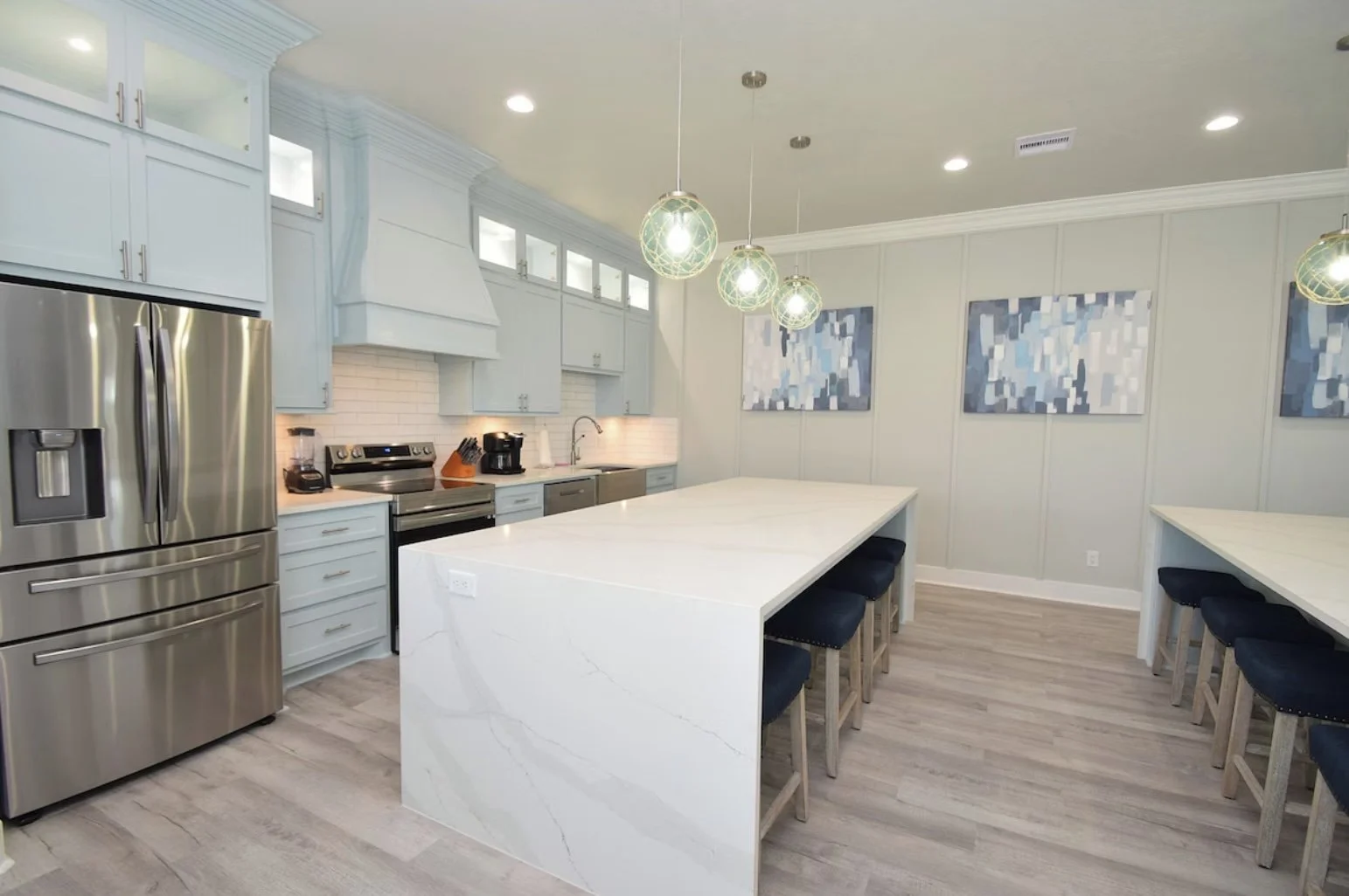 Modern kitchen with light blue cabinets, stainless steel refrigerator, white marble island, and artwork on the walls.