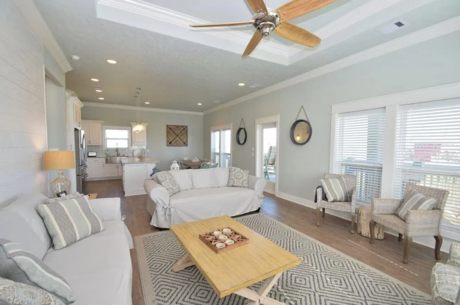 Living room with light-colored couches and chairs, wooden coffee table, patterned rug, large windows with blinds, ceiling fan, and open kitchen in the background.