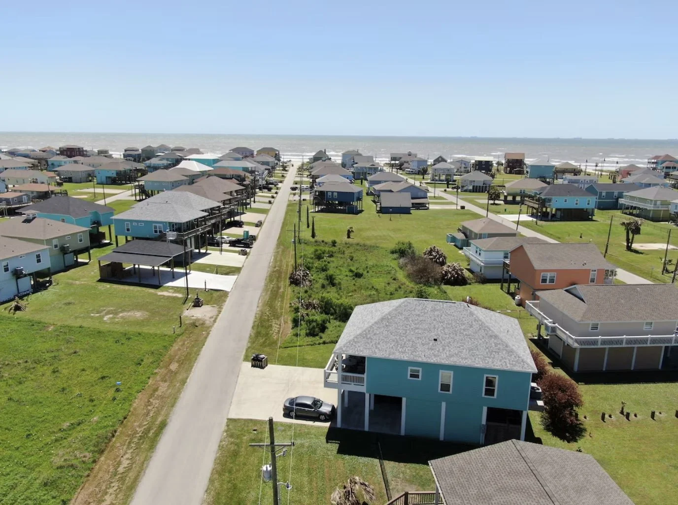Aerial view of a beachside neighborhood with colorful houses, green lawns, and a road leading to the ocean in the background.