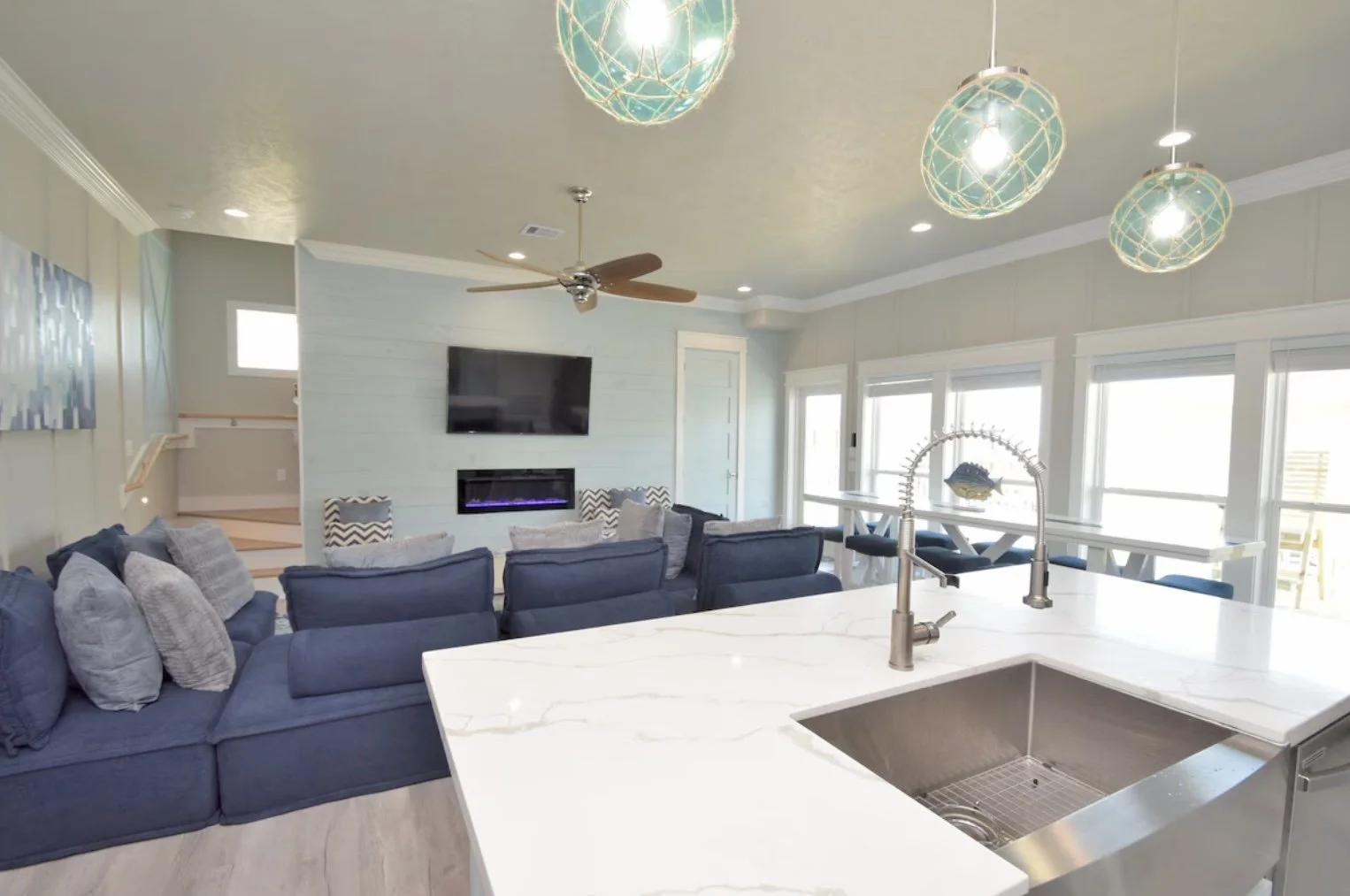 Open-concept living room and kitchen with blue sectional sofa, mounted TV, fireplace, ceiling fan, multiple pendant lights, white marble island with sink, and large windows.