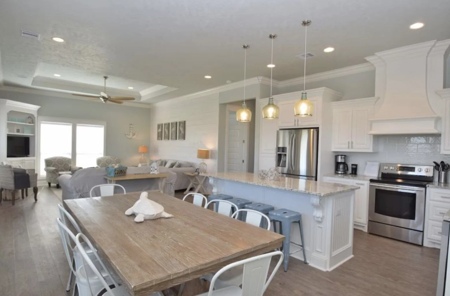 Open-concept kitchen and living room with white cabinetry, stainless steel appliances, a large wooden dining table with white chairs, and a cozy sitting area with armchairs and a sofa, decorated in a beach-inspired style.