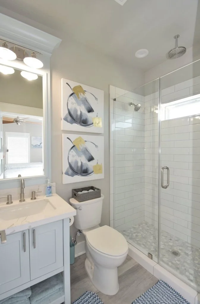 A modern bathroom with a glass-enclosed shower, a white vanity with a marble countertop, a mirror with lights, and two abstract art pieces on the wall. The bathroom also has a toilet with a small basket on top and a blue and white bath mat.