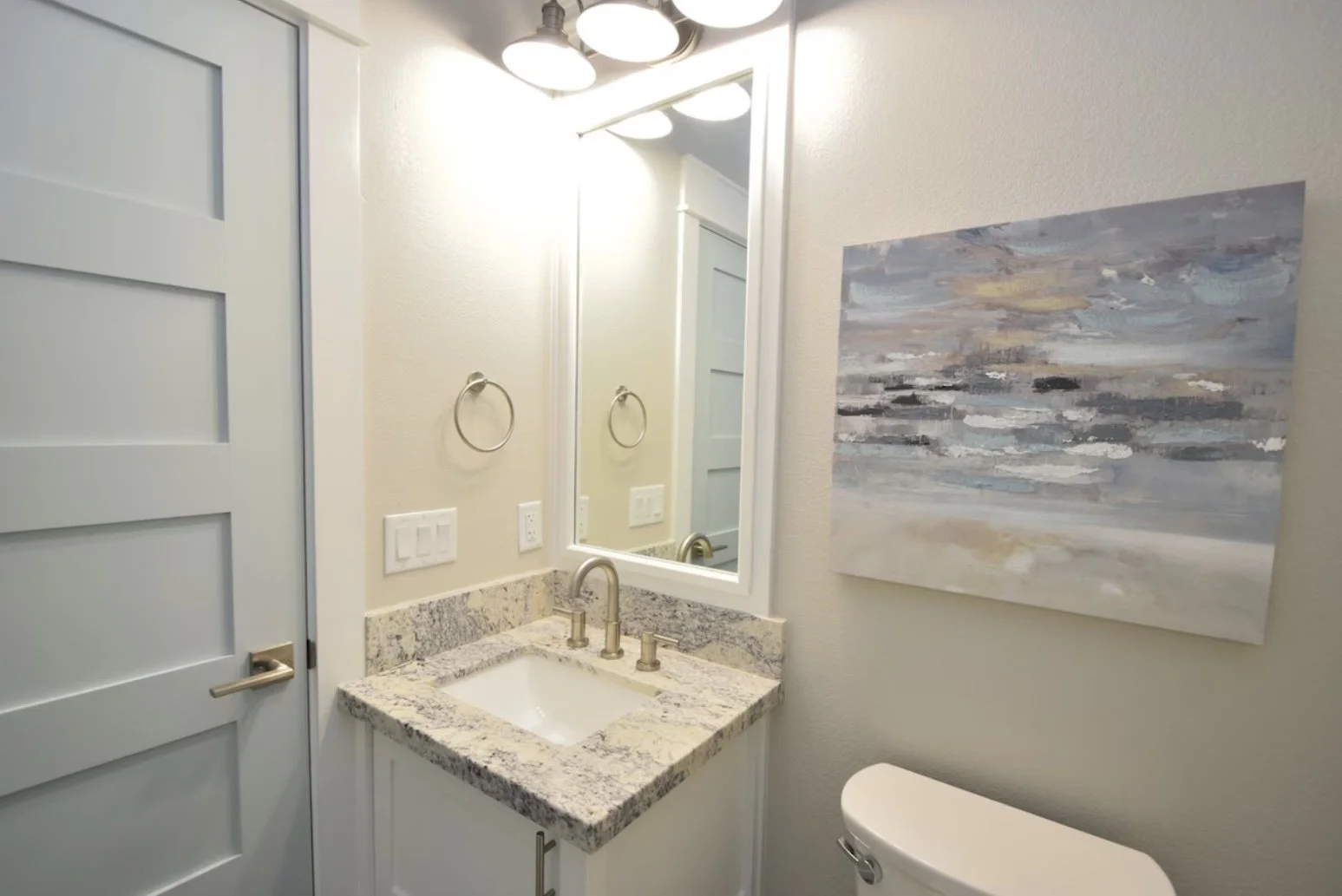 A small bathroom with a granite sink counter, a mirror, a wall-mounted towel ring, a light fixture above the mirror, a closed door, and abstract artwork on the wall.