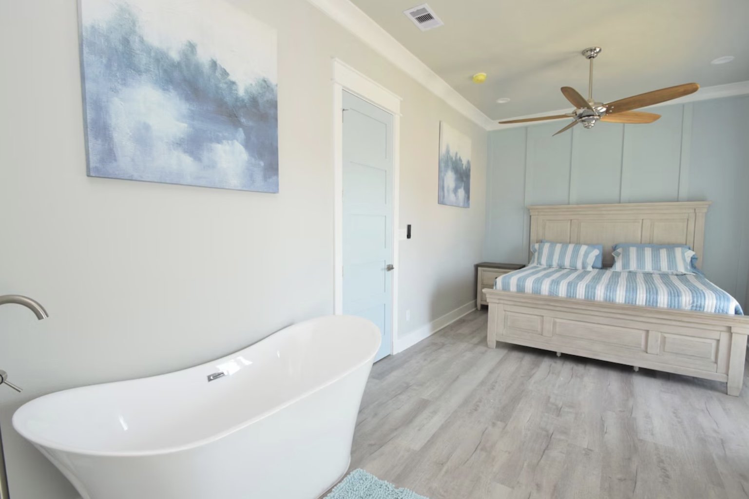 A bedroom with a wooden bed with blue and white striped bedding, an artwork featuring blue and white clouds, a corner bathtub, a ceiling fan, and light-colored wood flooring.