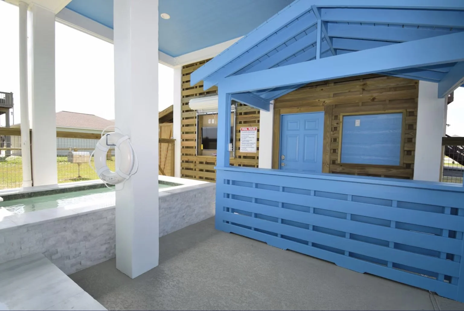 Outdoor pool area with a white pool with a rocky edge, a blue wooden structure resembling a small house, a flat-screen TV, a life preserver, and a sign with pool rules.