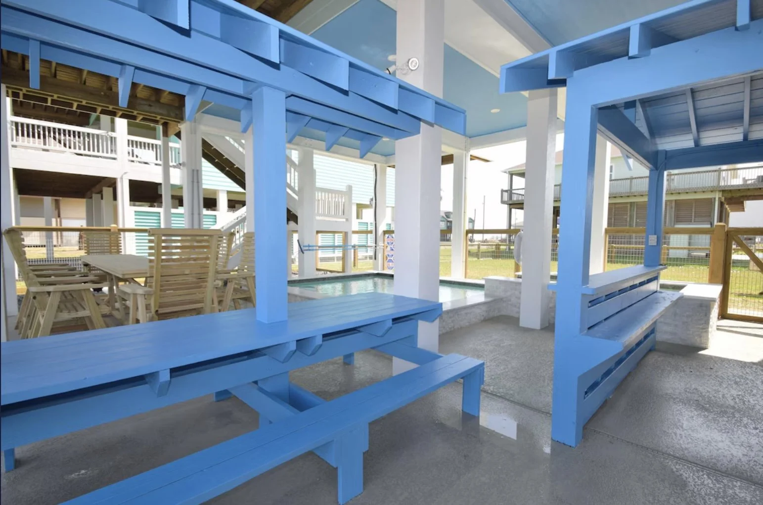 Outdoor covered seating area with blue wooden structure, benches, and a table, near a pool with a water slide in the background, at a beachside location.