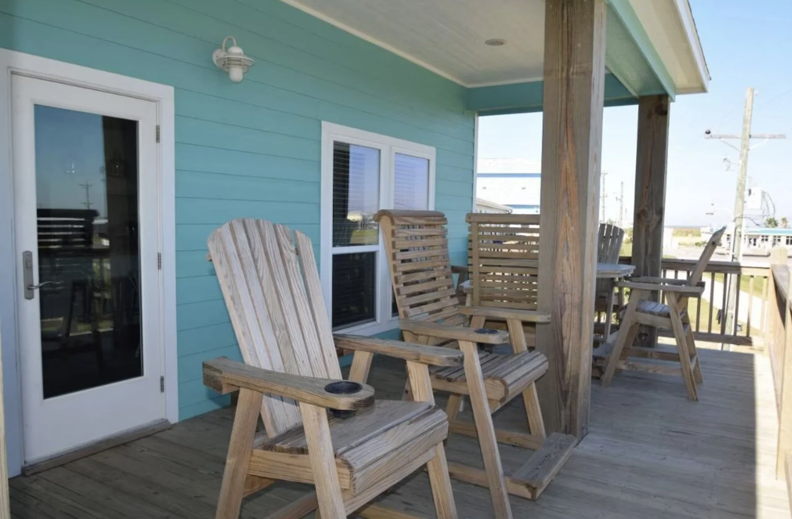 A wooden balcony with Adirondack chairs and a small table on a sunny day, attached to a turquoise house with white trim and a glass door.