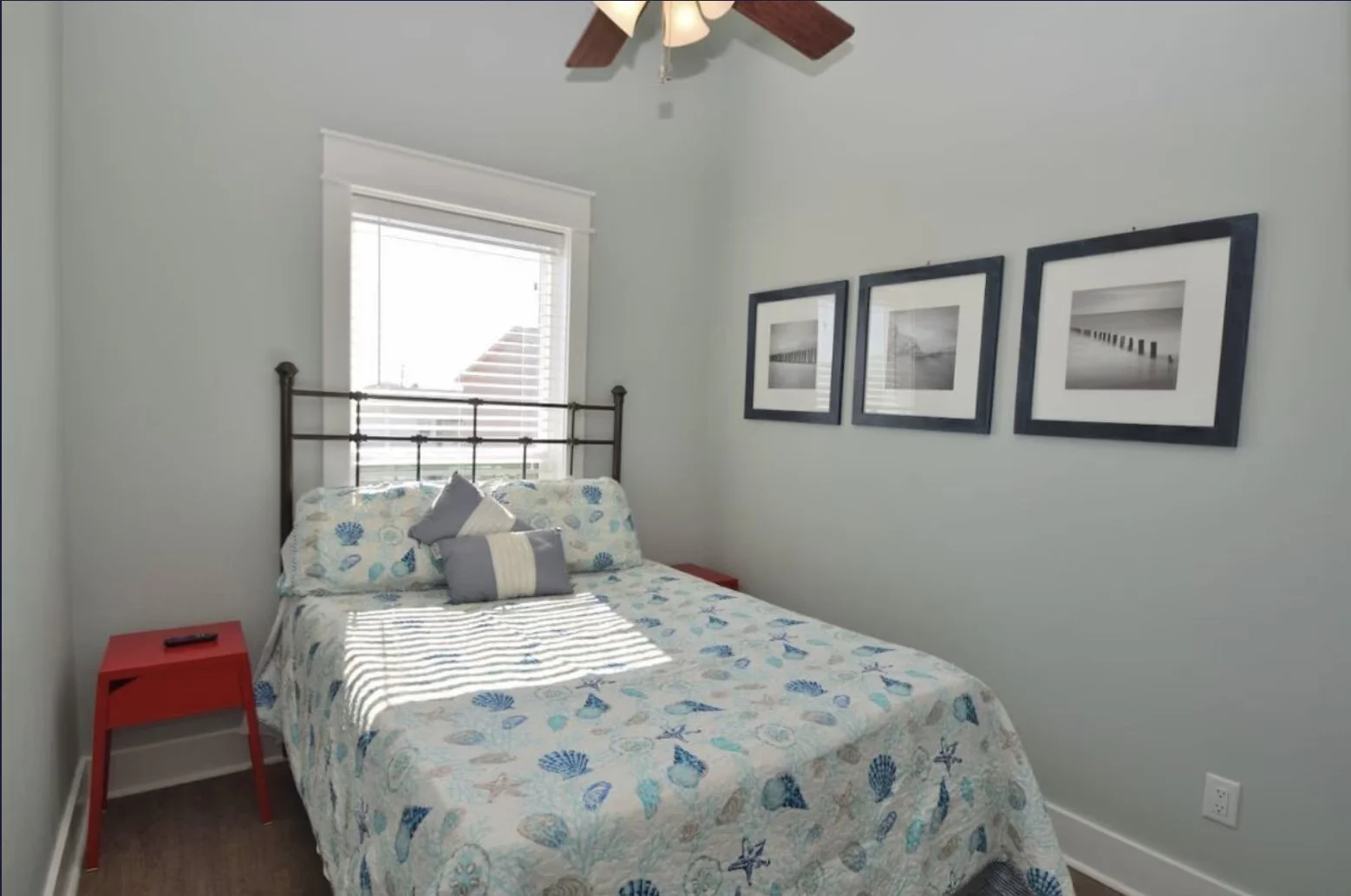 A bedroom with a bed covered in a seashell-themed bedspread, pillows, and a window with blinds, three black-framed photographs of a pier and bridge, a red side table, and a ceiling fan.