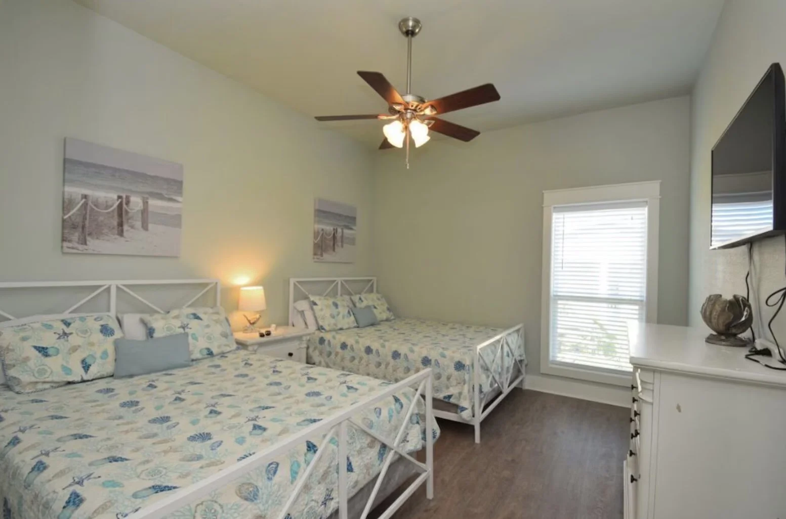 A bedroom with two white metal frame beds with beach-themed bedding, a nightstand with a lamp between the beds, wall art of a beach scene, a window with blinds, a ceiling fan with lights, and a white dresser.