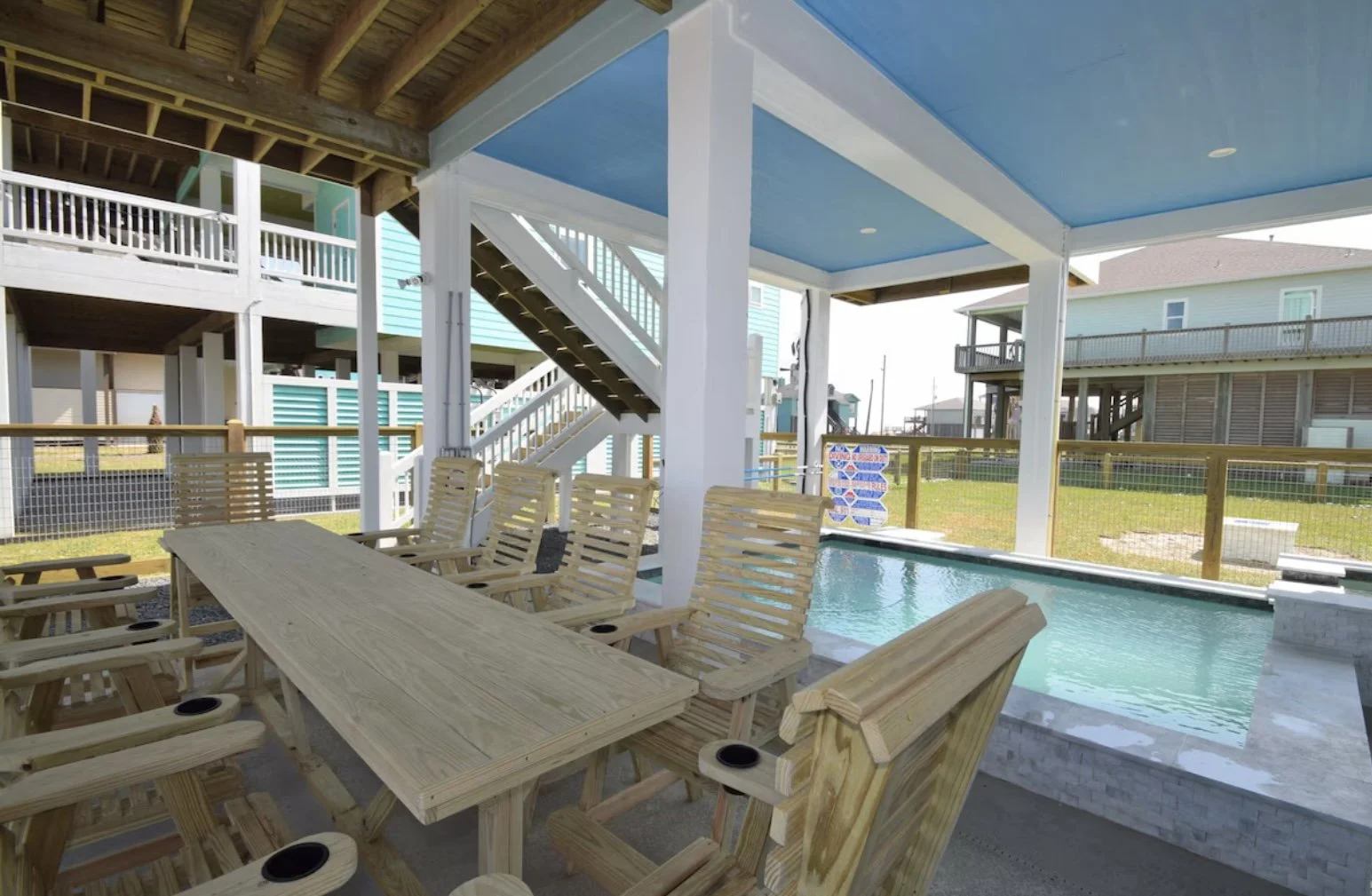 An outdoor patio area with wooden furniture, a table and chairs, overlooking a small pool. There are houses in the background, some with balconies, and a fence surrounds the patio, with clear skies above.