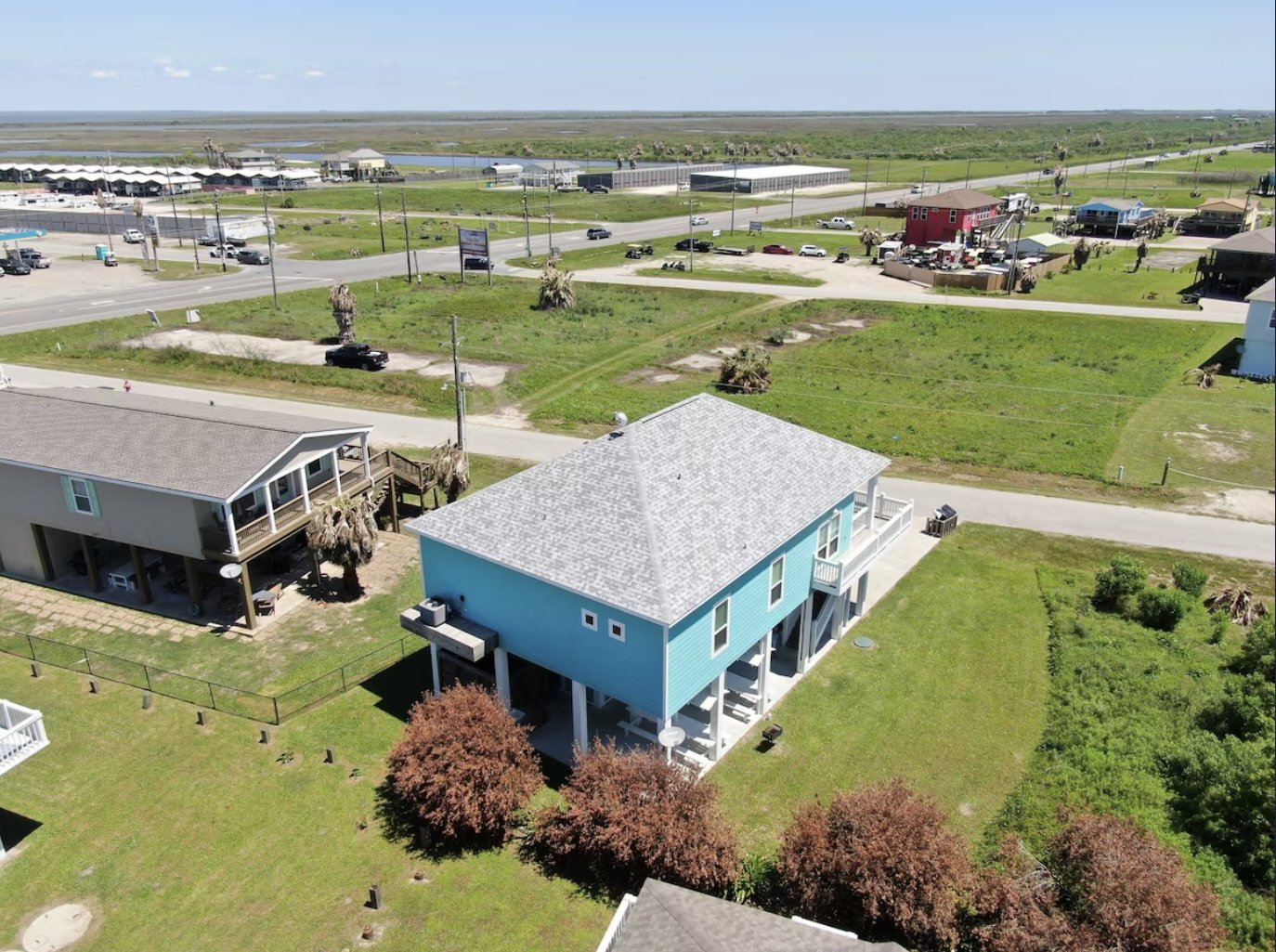 Aerial view of a coastal neighborhood with colorful houses, green lawns, palm trees, and a road with parked and moving cars. Water and marsh area in the background.