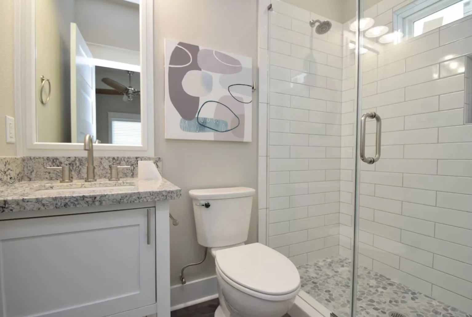 Bathroom with vanity, mirror, toilet, wall art, and glass-enclosed shower with pebble floor.