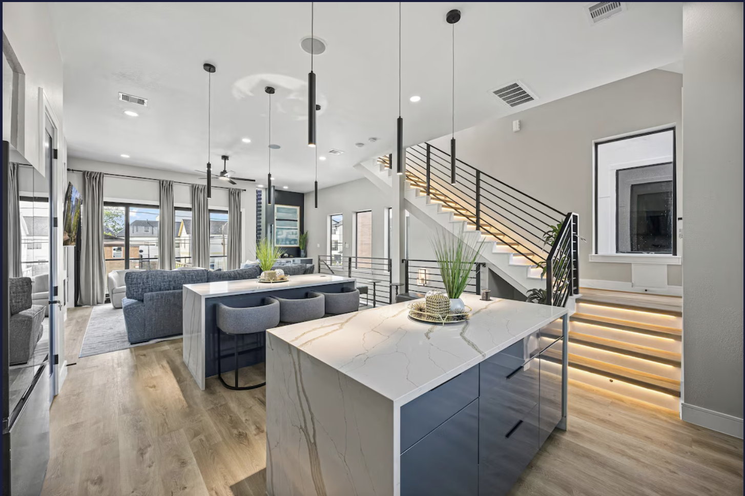 Interior of a modern open-concept living room and kitchen with marble island, gray cabinets, large windows, and staircase with black railing.