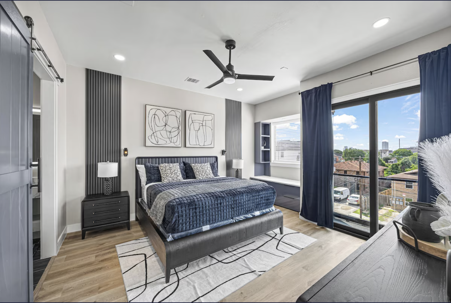 Modern bedroom with a large bed, black nightstand, white and black artwork, window seat, large sliding glass door with dark curtains, ceiling fan, and window view of a neighborhood.