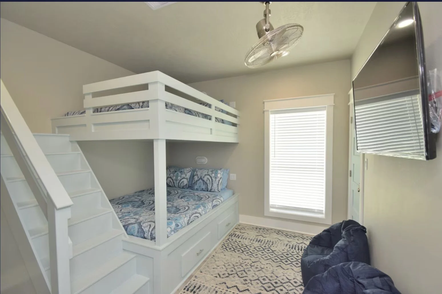 Bedroom with a lower and upper bunk bed, a window with blinds, a wall-mounted TV, and a patterned rug on the floor.