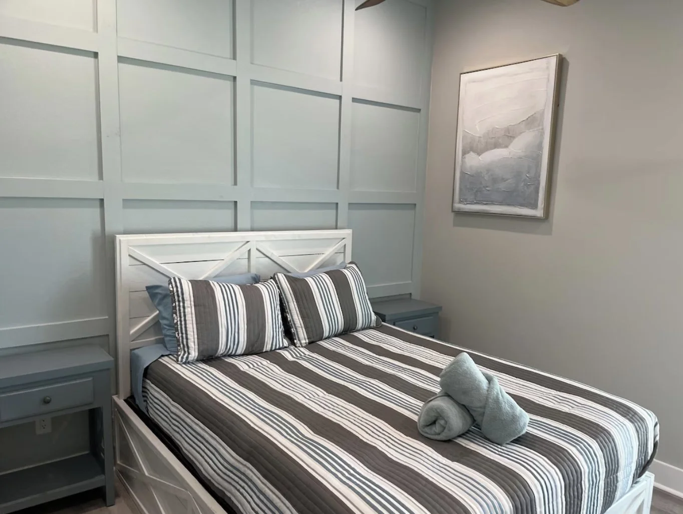A neatly made bed with striped black, white, and gray bedding, two matching pillows, and a rolled towel on top, in a bedroom with light gray paneled walls, a gray nightstand, and a framed abstract painting.