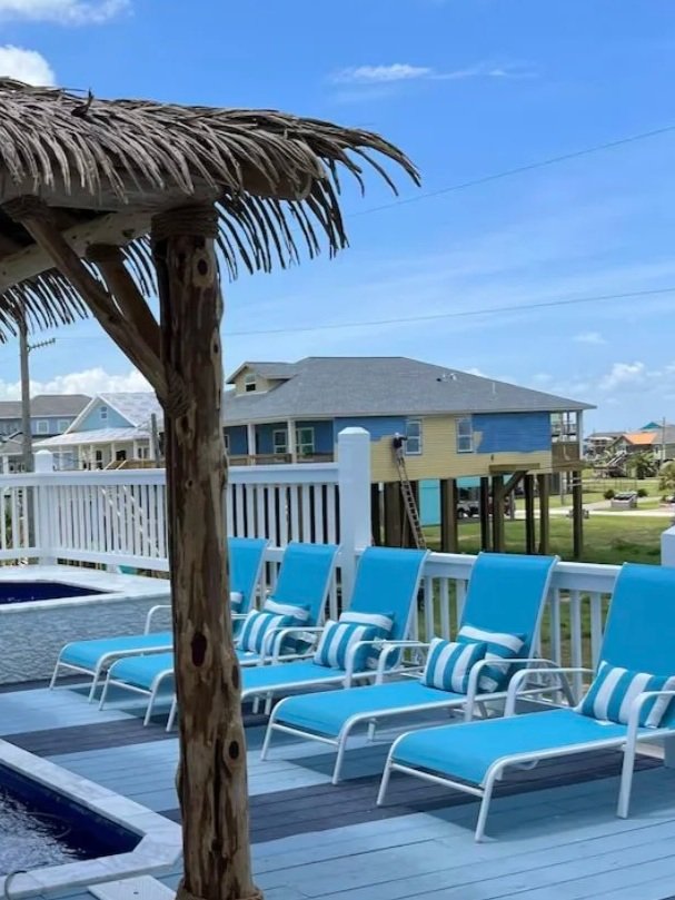 Outdoor pool area with four blue lounge chairs and striped pillows under a thatched umbrella, neighboring houses on stilts in the background, and a partly cloudy sky.