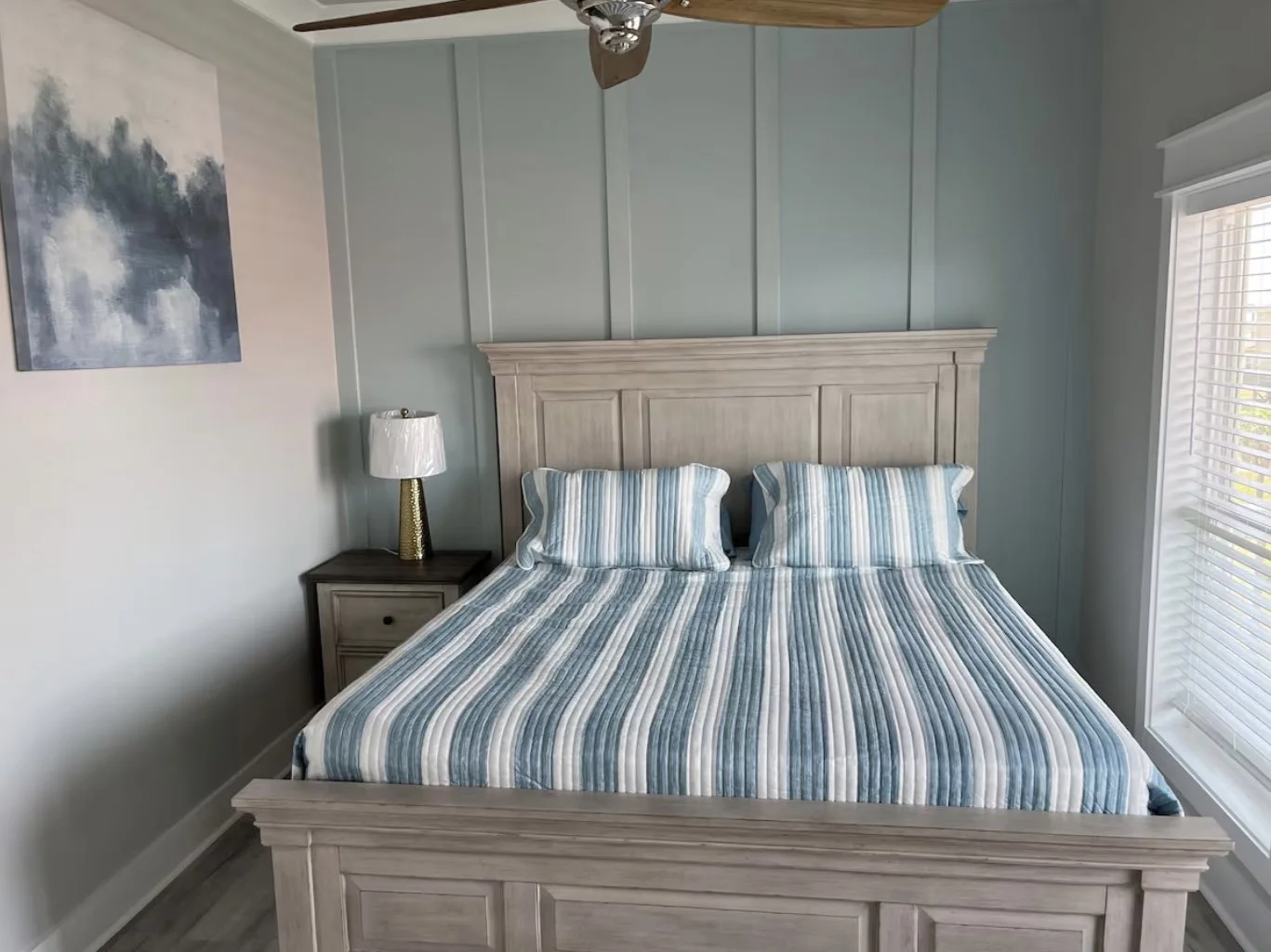 A bedroom with a light wood bed, striped blue and white bedding, and matching pillows. A nightstand with a gold lamp is beside the bed. There is a window with blinds on the right and a blue abstract painting on the left wall.