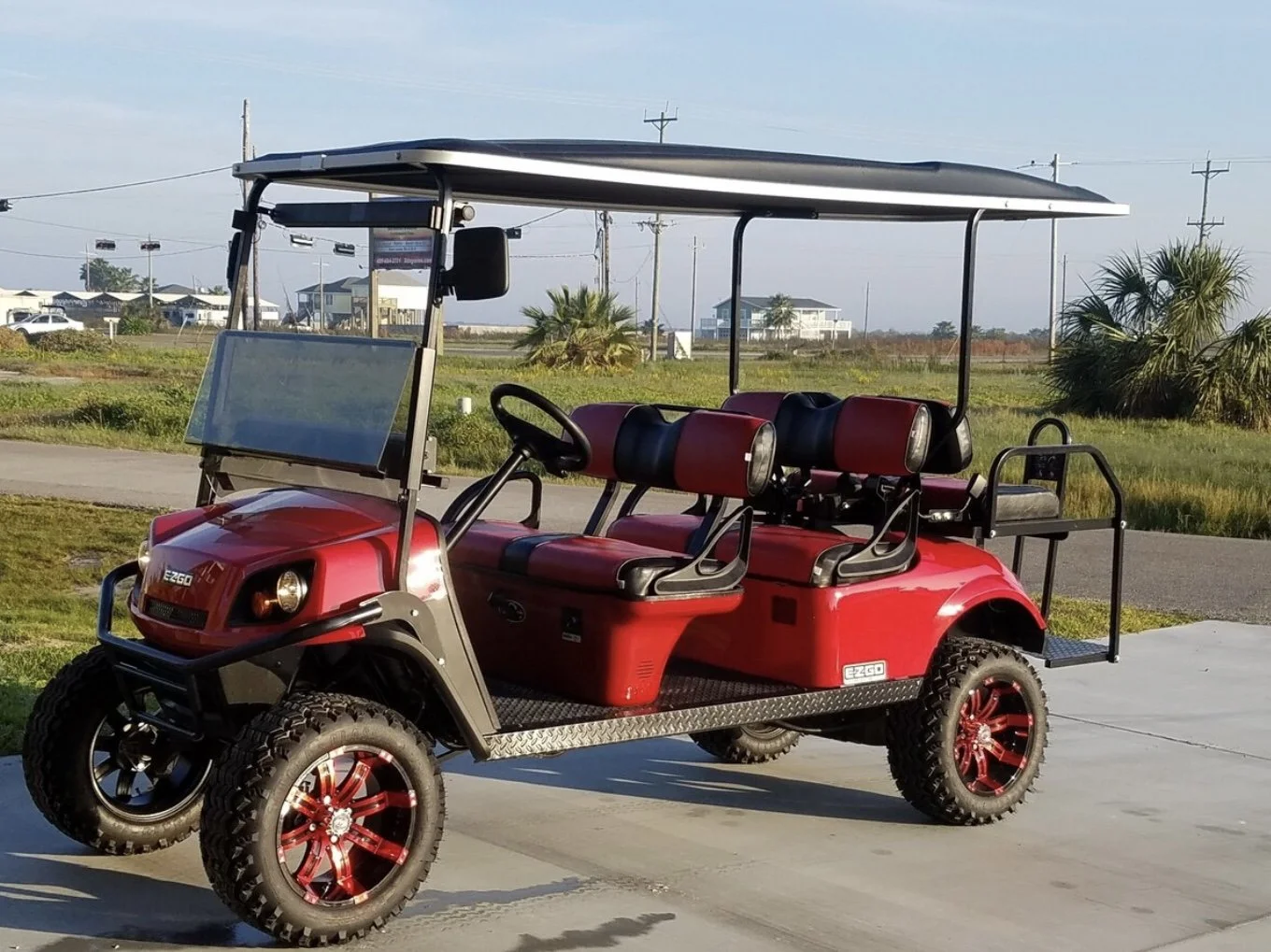 A red electric golf cart with four seats and large wheels parked on a paved surface in an open outdoor area with grass, palm trees, and buildings in the background.