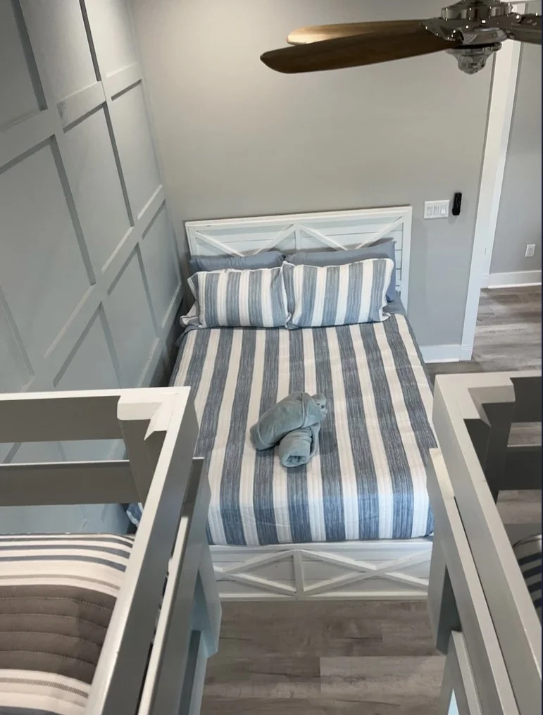 A cozy bedroom with a white wooden bed, striped bedding in shades of blue, gray, and white, and a rolled towel on the bed. The room has a ceiling fan with wooden blades, gray paneled walls, and gray flooring.