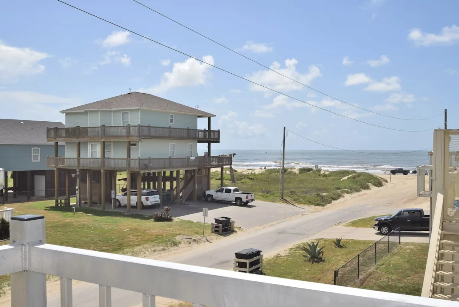A beachside scene with a blue multi-story house on stilts, parked cars, a sandy road, green bushes, and the ocean in the background under a blue sky with scattered clouds.
