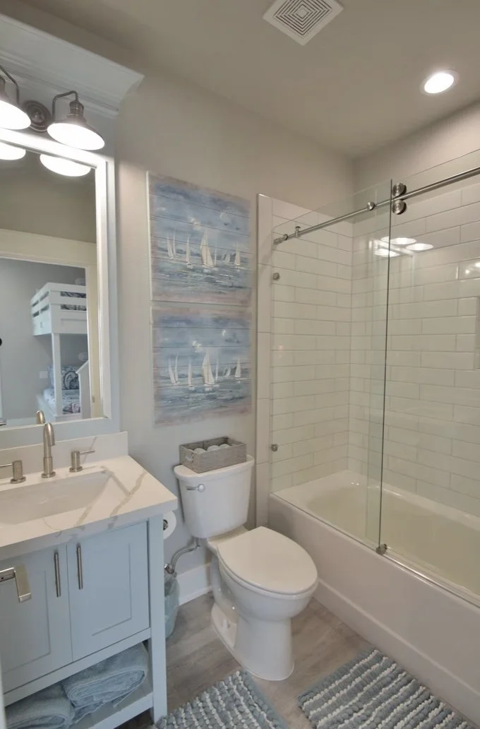 Modern bathroom with a white vanity, a toilet, and a glass-enclosed bathtub with white subway tiles. Decor includes boat-themed wall art and striped rugs.