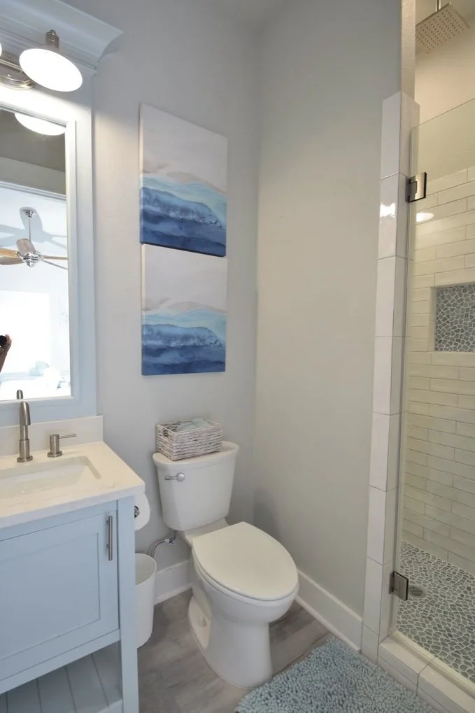 Bathroom with a blue and white abstract painting, a white vanity with a mirror, a toilet with a basket on top, and a tiled shower stall with glass door.