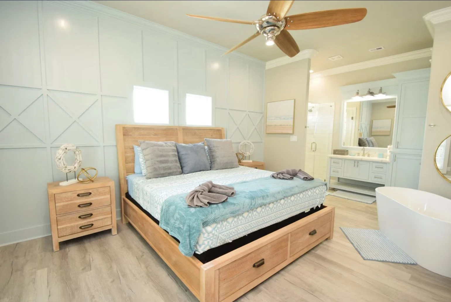 A cozy bedroom with a wooden bed, gray pillows, and a turquoise blanket. A wooden nightstand with decorative items is next to the bed. There is a ceiling fan with wooden blades and windows behind the bed. An attached bathroom with a large mirror, sin