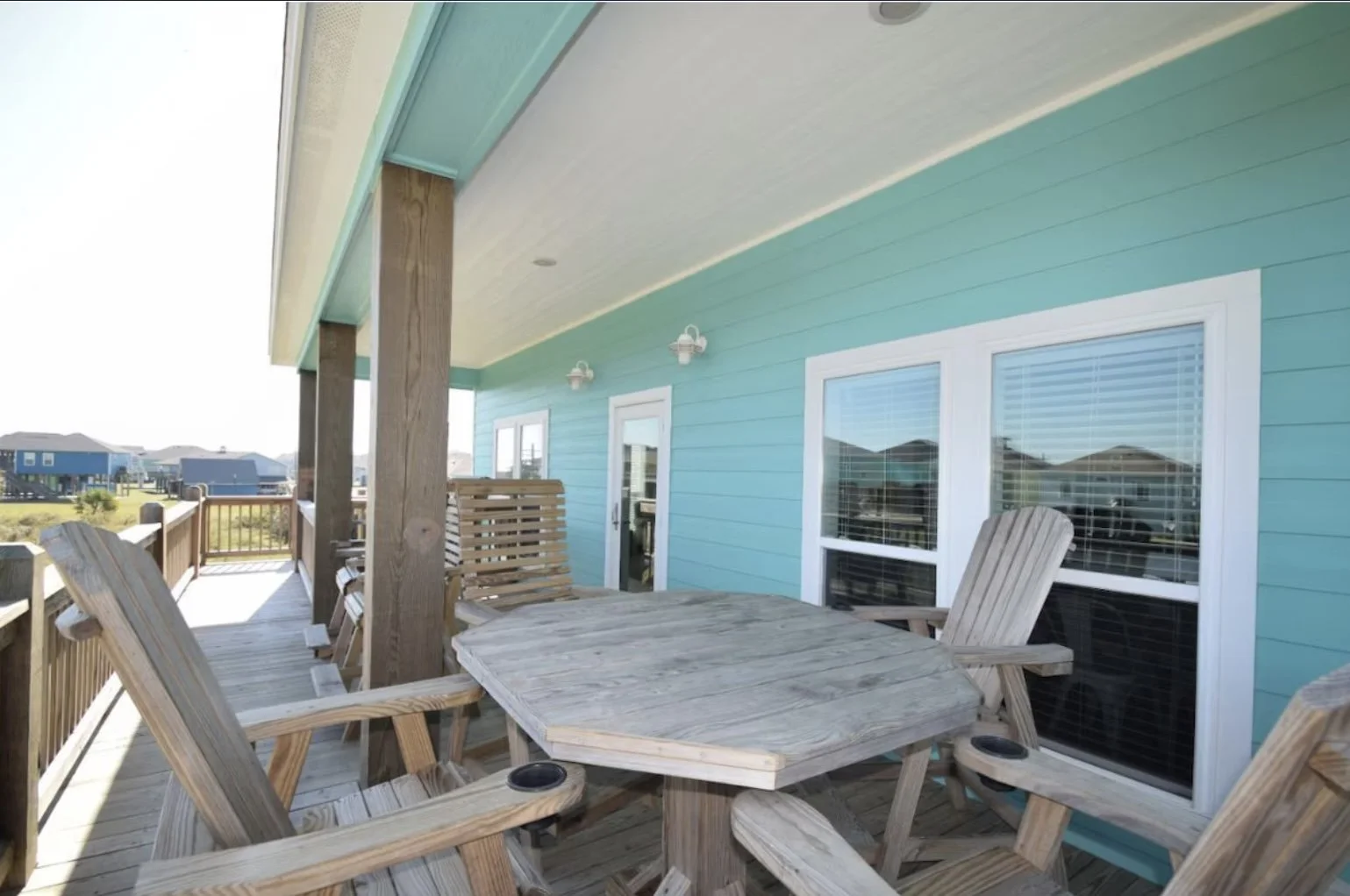 Back porch with wooden table and chairs, teal siding, white window frames, outdoor lighting, and neighboring houses in the background.
