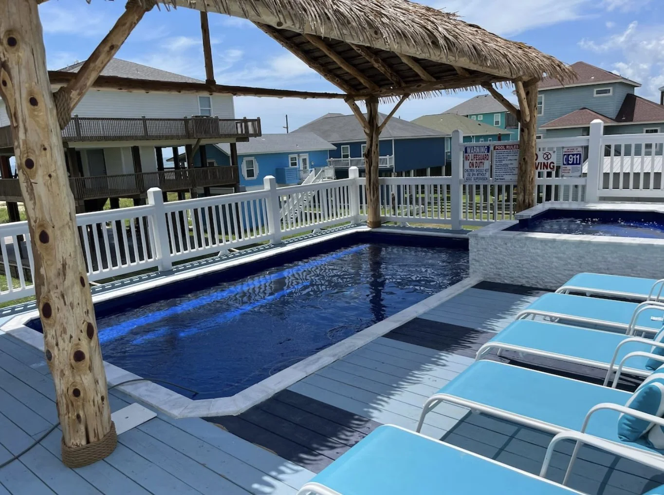 A backyard pool area with a hot tub, white fencing, and a thatched roof cabana. Lounge chairs with blue cushions are lined up on the deck, which has a wooden floor and nearby residential houses in the background.