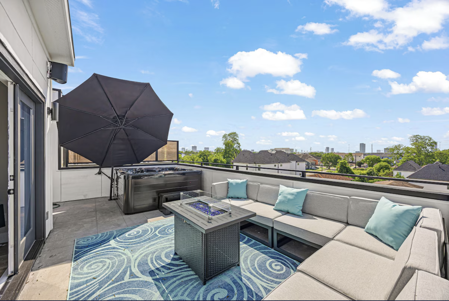 Rooftop patio with sectional sofa, blue pillows, rectangular table, large black umbrella, and view of city skyline under blue sky with scattered clouds.